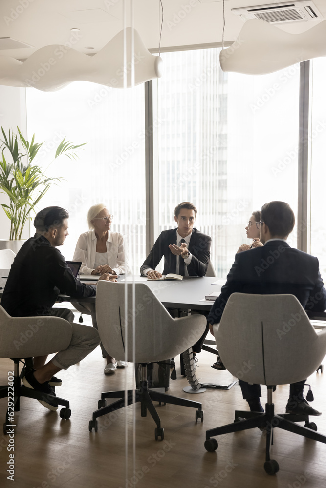 Obraz premium Young boss, CEO, executive speaking to managers on team meeting, sitting at conference table, discussing project management, plan, strategy. Business investors negotiating on deal. Through glass view