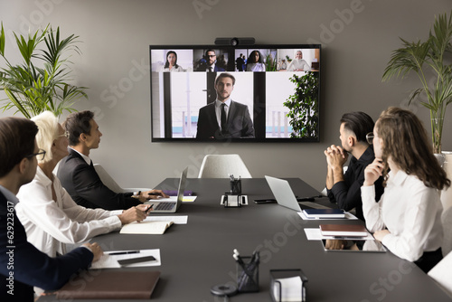 Business team and leader meeting online and offline, sitting at conference table, looking at interactive board with headshots, listening to speaker, CEO, discussing project on group video call