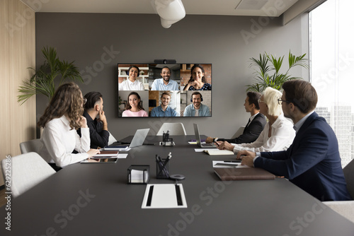 Office staff meeting with diverse freelance team on online video conference, making group call on internet, sitting at table looking at electronic board with head shots, discussing work project