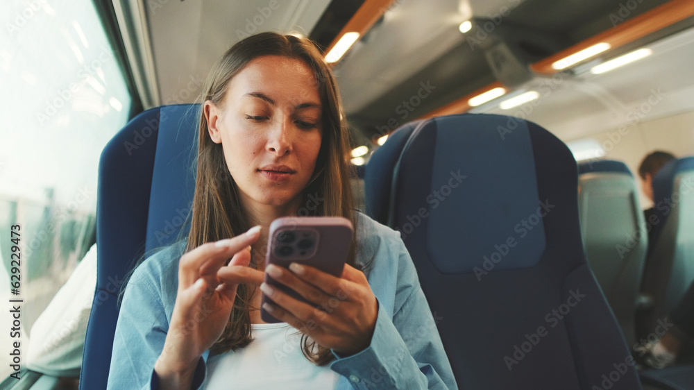 Cute girl with long brown hair, dressed in blue shirt, looks out window while traveling by suburban train, scrolls on smartphone screen
