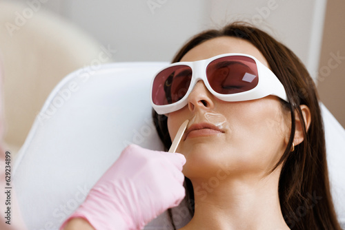 Closeup of beautician hand using wooden spatula for applying transparent anaesthetic gel on woman's face for making the laser removal hair procedure. Laser hair removal photo