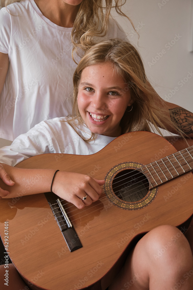 Fototapeta premium Portrait of a blonde girl playing guitar while smiling on camera with a woman sitting with he