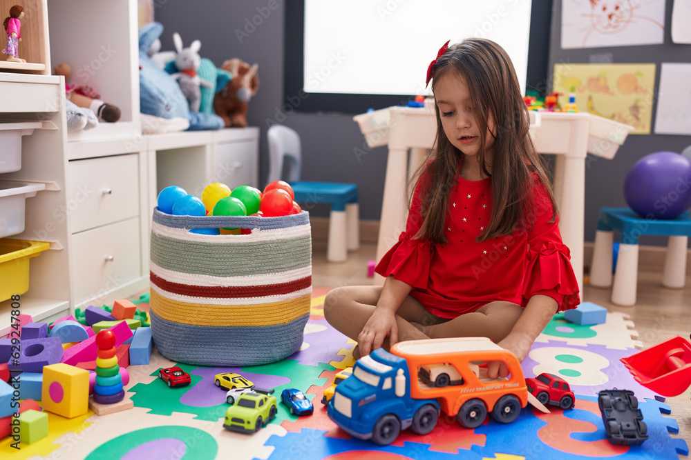 Fototapeta premium Adorable hispanic girl playing with car toy sitting on floor at kindergarten