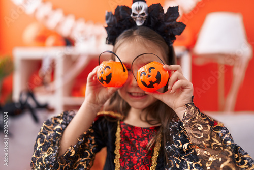 Adorable hispanic girl wearing halloween costume covering eyes with small pumpkin baskets at home photo