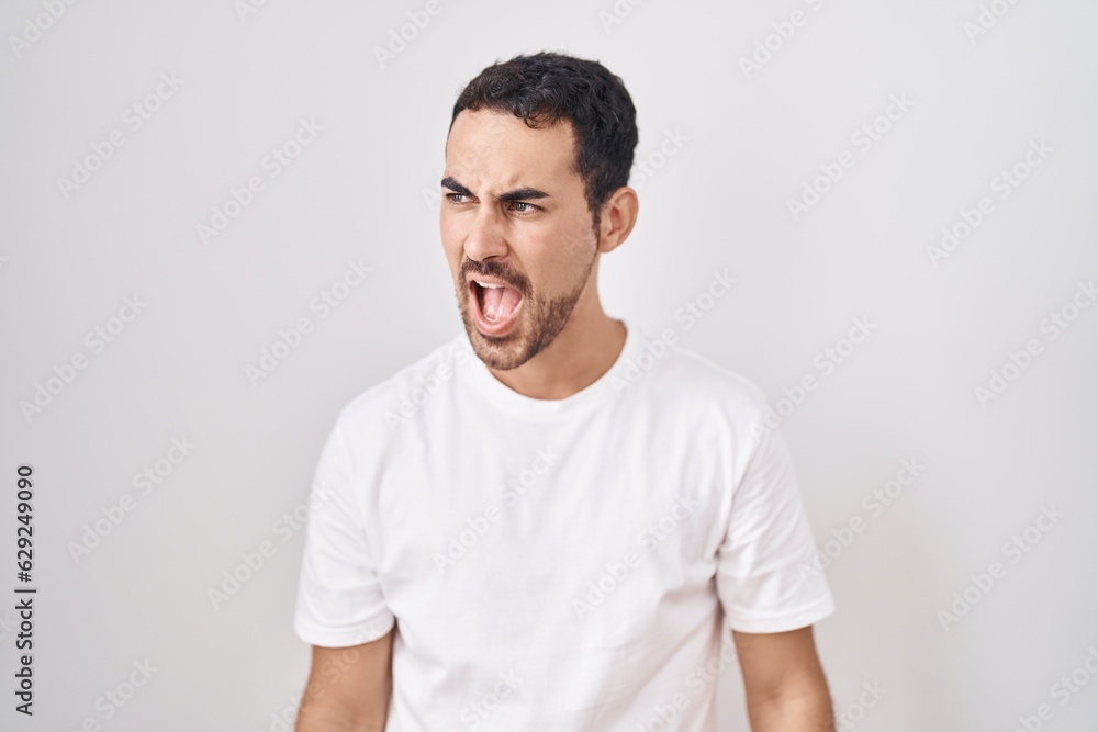 Handsome hispanic man standing over white background angry and mad screaming frustrated and furious, shouting with anger. rage and aggressive concept. Handsome hispanic man standing over white background angry and mad screaming frustrated and furious, shouting with anger. rage and aggressive concept.