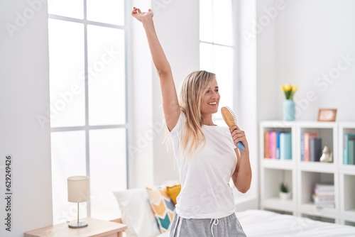 Young blonde woman singing song using brush as a microphone at bedroom