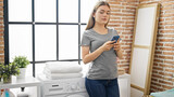 Young blonde woman using smartphone waiting for washing machine at laundry room