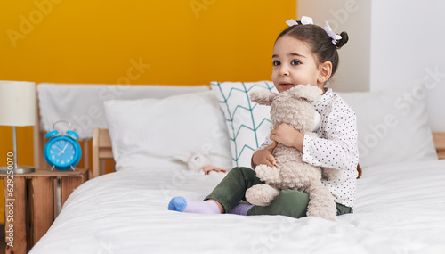 Adorable hispanic girl hugging teddy bear sitting on bed at bedroom