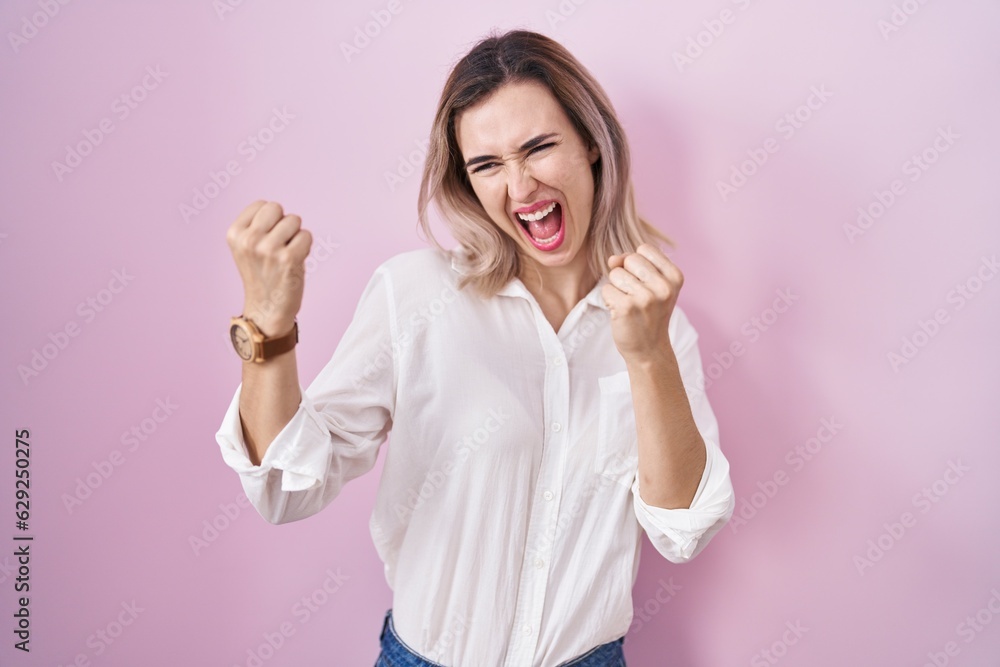 Young beautiful woman standing over pink background very happy and excited doing winner gesture with arms raised, smiling and screaming for success. celebration concept. Young beautiful woman standing over pink background very happy and excited doing winner gesture with arms raised, smiling and screaming for success. celebration concept.