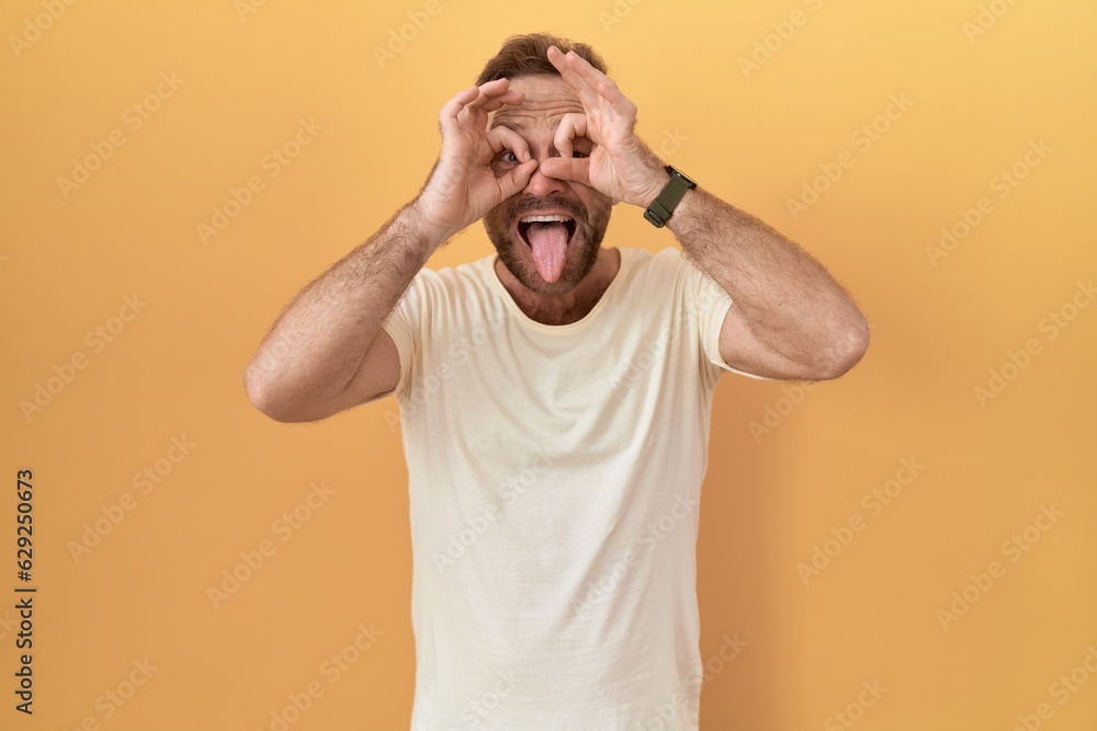 Middle age man with beard standing over yellow background doing ok gesture like binoculars sticking tongue out, eyes looking through fingers. crazy expression. Middle age man with beard standing over yellow background doing ok gesture like binoculars sticking tongue out, eyes looking through fingers. crazy expression.
