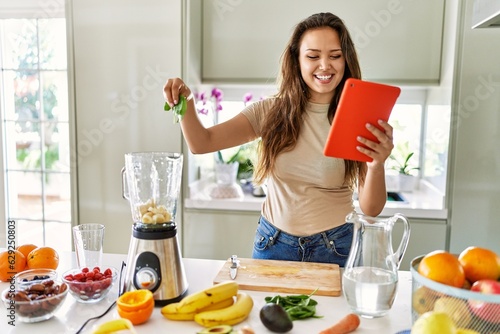Young beautiful hispanic woman preparing smoothie with blender looking at recipe on tablet at the kitchen