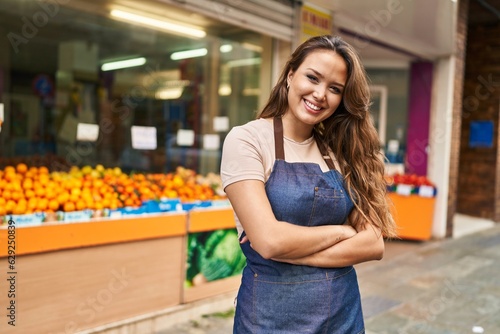 Young beautiful hispanic woman waitress smiling confident standing with arms crossed gesture at fruit store