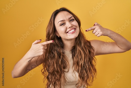 Young hispanic woman standing over yellow background smiling cheerful showing and pointing with fingers teeth and mouth. dental health concept.