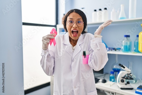 Young hispanic doctor woman working at scientist laboratory holding pink ribbon celebrating victory with happy smile and winner expression with raised hands