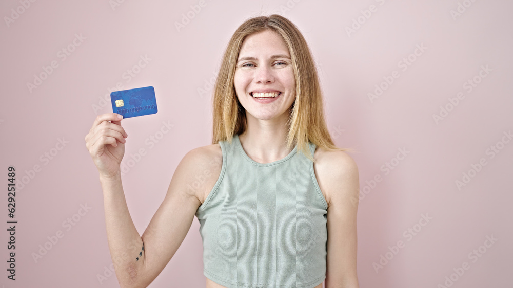 Young blonde woman smiling confident holding credit card over isolated pink background Young blonde woman smiling confident holding credit card over isolated pink background