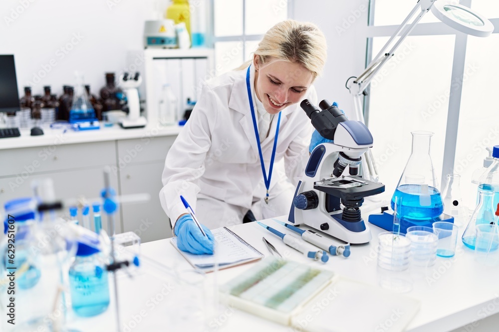 Young blonde woman scientist using microscope taking notes at laboratory Young blonde woman scientist using microscope taking notes at laboratory