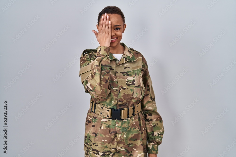 Beautiful african american woman wearing camouflage army uniform covering one eye with hand, confident smile on face and surprise emotion.