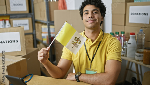 Young latin man volunteer holding vatican city flag smiling at charity center