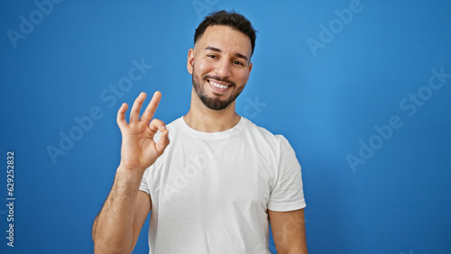 Young arab man smiling confident doing ok sign over isolated blue background