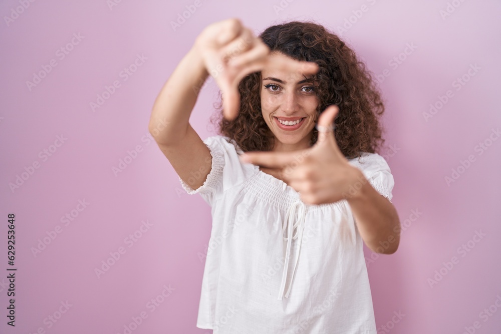 Hispanic woman with curly hair standing over pink background smiling making frame with hands and fingers with happy face. creativity and photography concept. Hispanic woman with curly hair standing over pink background smiling making frame with hands and fingers with happy face. creativity and photography concept.