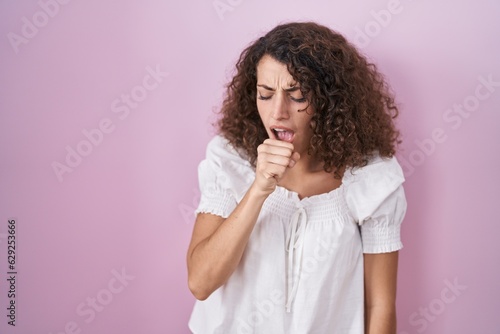 Hispanic woman with curly hair standing over pink background feeling unwell and coughing as symptom for cold or bronchitis. health care concept.