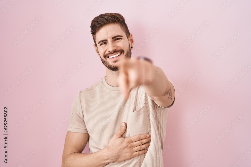 Hispanic man with beard standing over pink background laughing at you, pointing finger to the camera with hand over body, shame expression Hispanic man with beard standing over pink background laughing at you, pointing finger to the camera with hand over body, shame expression