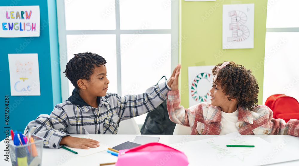 Naklejka premium Adorable african american boy and girl students drawing on notebook high five at classroom