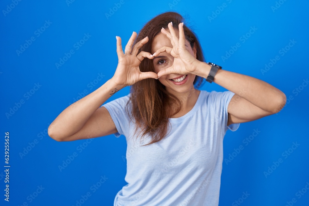 Brunette woman standing over blue background doing heart shape with hand and fingers smiling looking through sign Brunette woman standing over blue background doing heart shape with hand and fingers smiling looking through sign