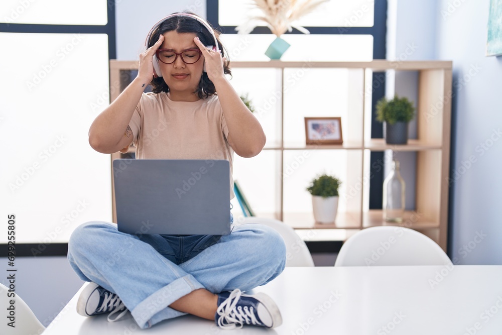 Young hispanic woman using laptop sitting on the table wearing headphones with hand on head for pain in head because stress. suffering migraine. Young hispanic woman using laptop sitting on the table wearing headphones with hand on head for pain in head because stress. suffering migraine.