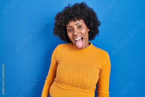Photography Black woman with curly hair standing over blue background sticking tongue out happy with funny expression