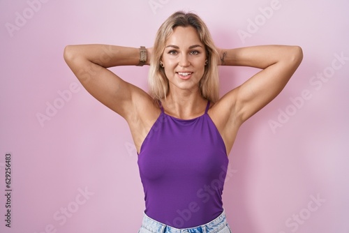 Young blonde woman standing over pink background relaxing and stretching  arms and hands behind head and neck smiling happy