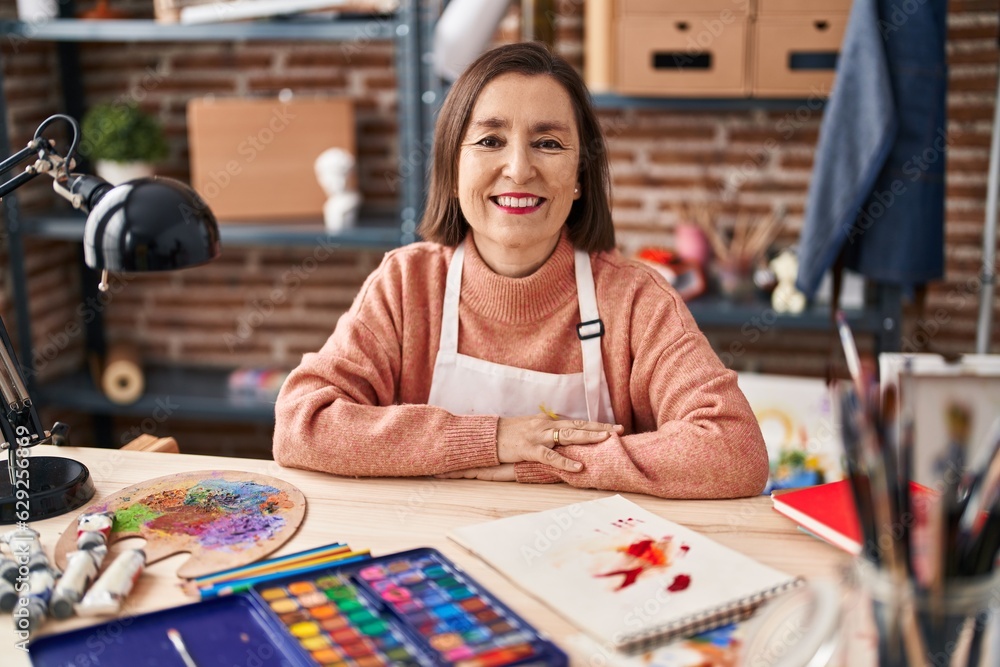 Middle age woman artist smiling confident sitting on table at art studio Middle age woman artist smiling confident sitting on table at art studio