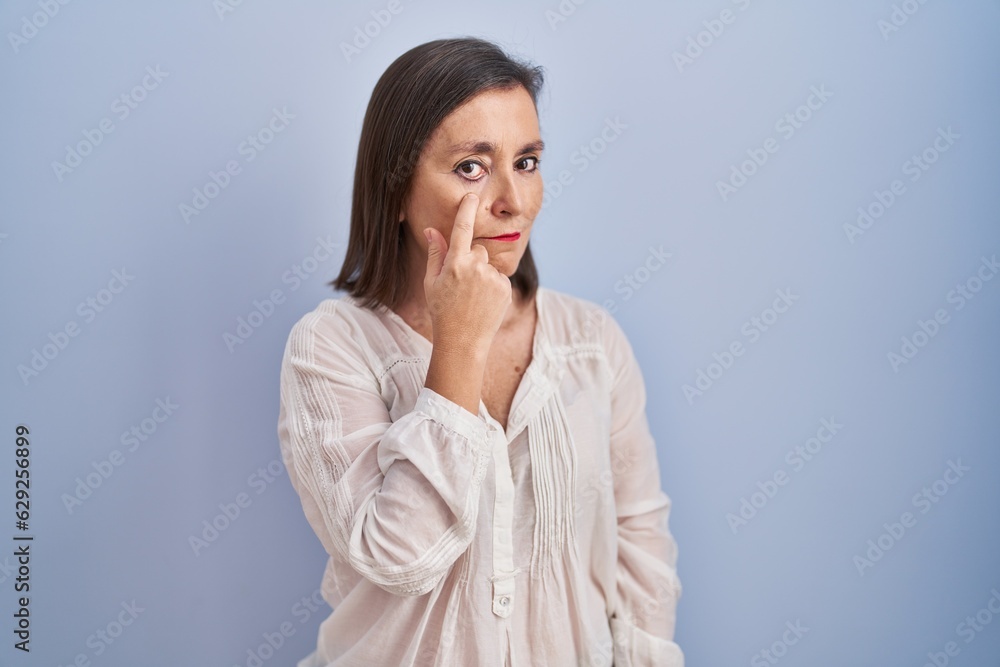 Middle age hispanic woman standing over blue background pointing to the eye watching you gesture, suspicious expression Middle age hispanic woman standing over blue background pointing to the eye watching you gesture, suspicious expression