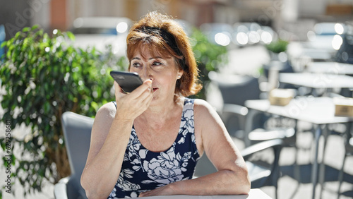 Fototapeta Naklejka Na Ścianę i Meble -  Middle age woman sending voice message by smartphone sitting on table at coffee shop terrace