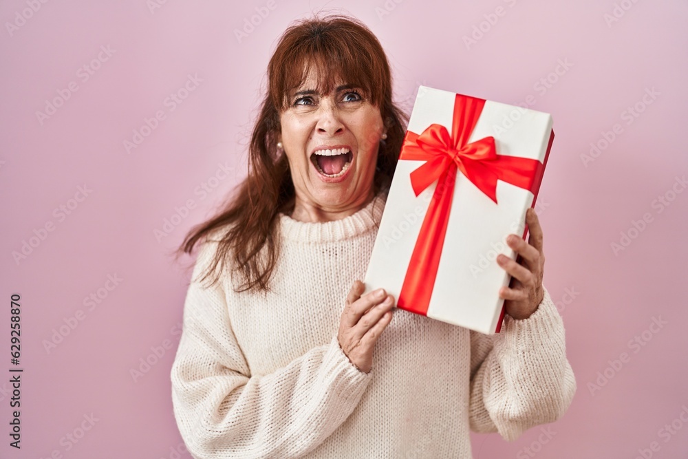 Middle age hispanic woman holding present angry and mad screaming frustrated and furious, shouting with anger looking up.