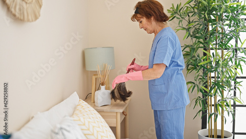 Middle age woman professional cleaner cleaning using duster at bedroom photo