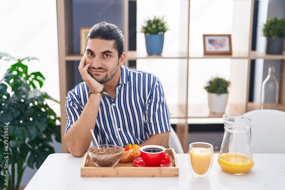 Hispanic man with long hair sitting on the table having breakfast thinking looking tired and bored with depression problems with crossed arms. Hispanic man with long hair sitting on the table having breakfast thinking looking tired and bored with depression problems with crossed arms.