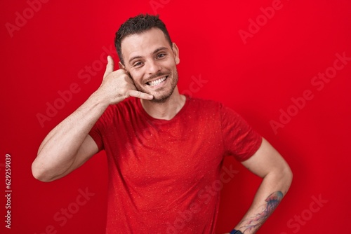 Young hispanic man standing over red background smiling doing phone gesture with hand and fingers like talking on the telephone. communicating concepts. © Krakenimages.com