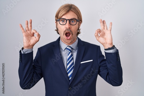 Caucasian man with mustache wearing business clothes looking surprised and shocked doing ok approval symbol with fingers. crazy expression