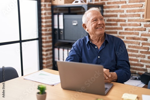 Senior man with grey hair working using computer laptop at the office looking away to side with smile on face, natural expression. laughing confident.