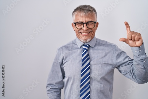 Hispanic business man with grey hair wearing glasses smiling and confident gesturing with hand doing small size sign with fingers looking and the camera. measure concept.