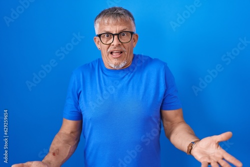 Hispanic man with grey hair standing over blue background clueless and confused with open arms, no idea concept.