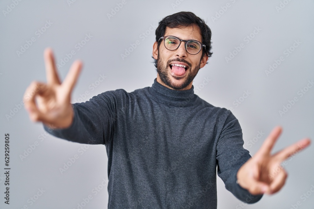 Handsome latin man standing over isolated background smiling with tongue out showing fingers of both hands doing victory sign. number two. Handsome latin man standing over isolated background smiling with tongue out showing fingers of both hands doing victory sign. number two.