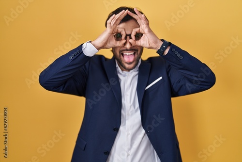 Handsome latin man standing over yellow background doing ok gesture like binoculars sticking tongue out, eyes looking through fingers. crazy expression.