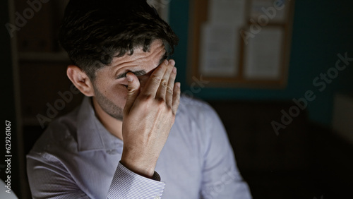 Young hispanic man business worker stressed sitting on table at the office
