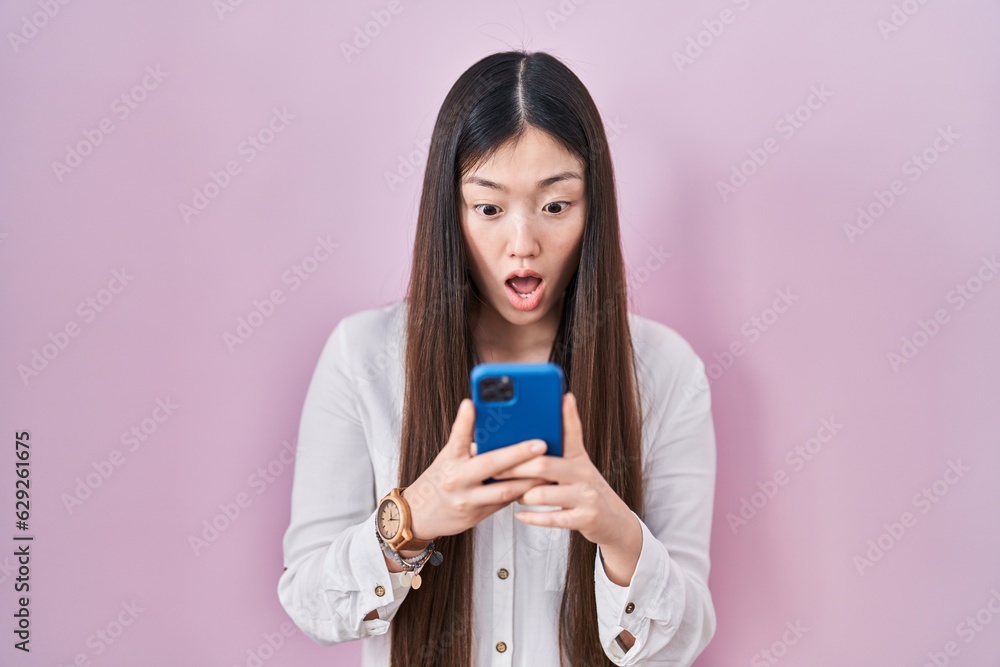 Chinese young woman using smartphone typing message afraid and shocked with surprise and amazed expression, fear and excited face.