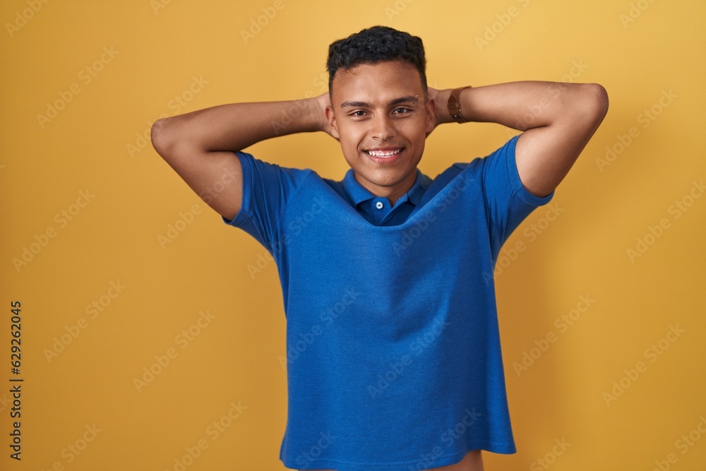 Young hispanic man standing over yellow background relaxing and stretching, arms and hands behind head and neck smiling happy Young hispanic man standing over yellow background relaxing and stretching, arms and hands behind head and neck smiling happy