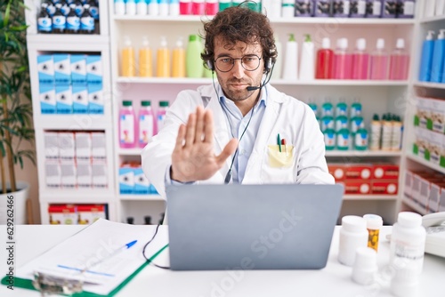Hispanic young man working at pharmacy drugstore working with laptop with open hand doing stop sign with serious and confident expression, defense gesture