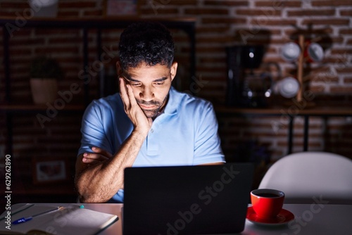 Hispanic man with beard using laptop at night thinking looking tired and bored with depression problems with crossed arms.