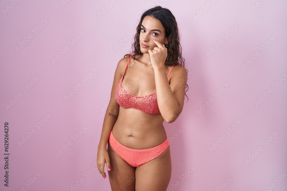 Naklejka premium Young hispanic woman wearing lingerie over pink background pointing to the eye watching you gesture, suspicious expression